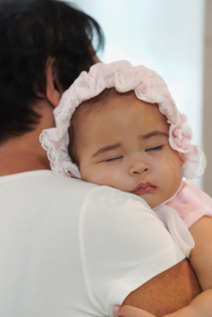 A baby in a bonnet asleep on a grown-up's shoulder.