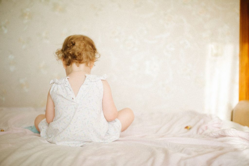 A baby sitting up on a bed, seen from behind.