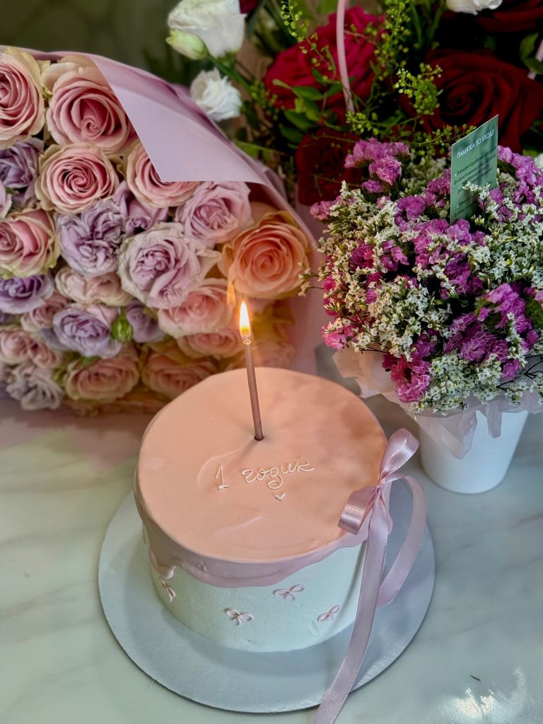 A pink birthday cake with one candle and flowers.