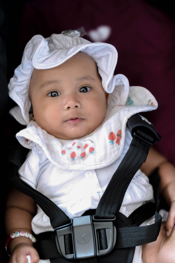 A baby in a white hat, buckled into a car seat.