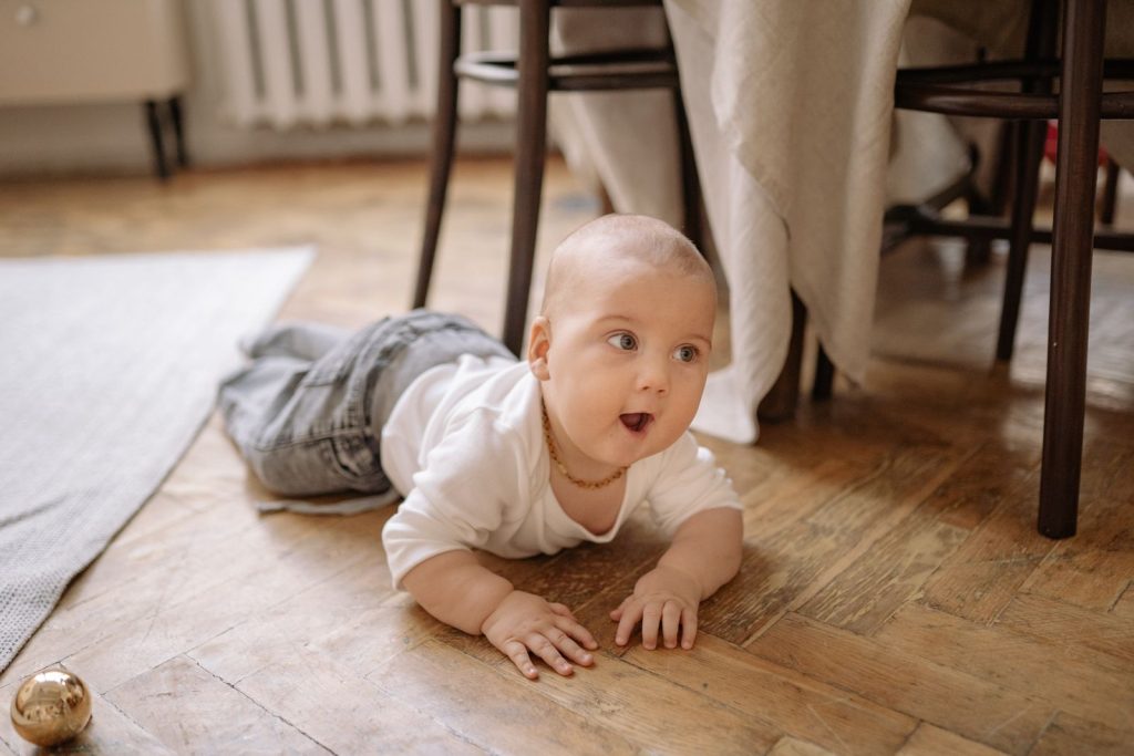 A baby in white crawling on a wooden floor.