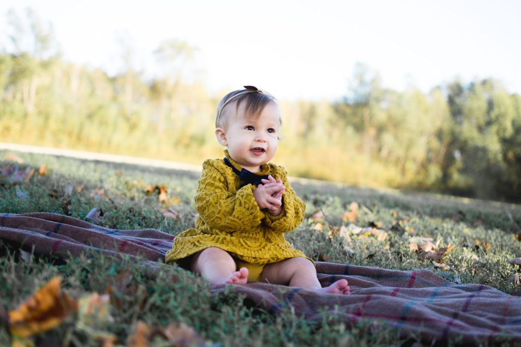 A smiling baby girl sitting on a blanket outdoors in the fall.
